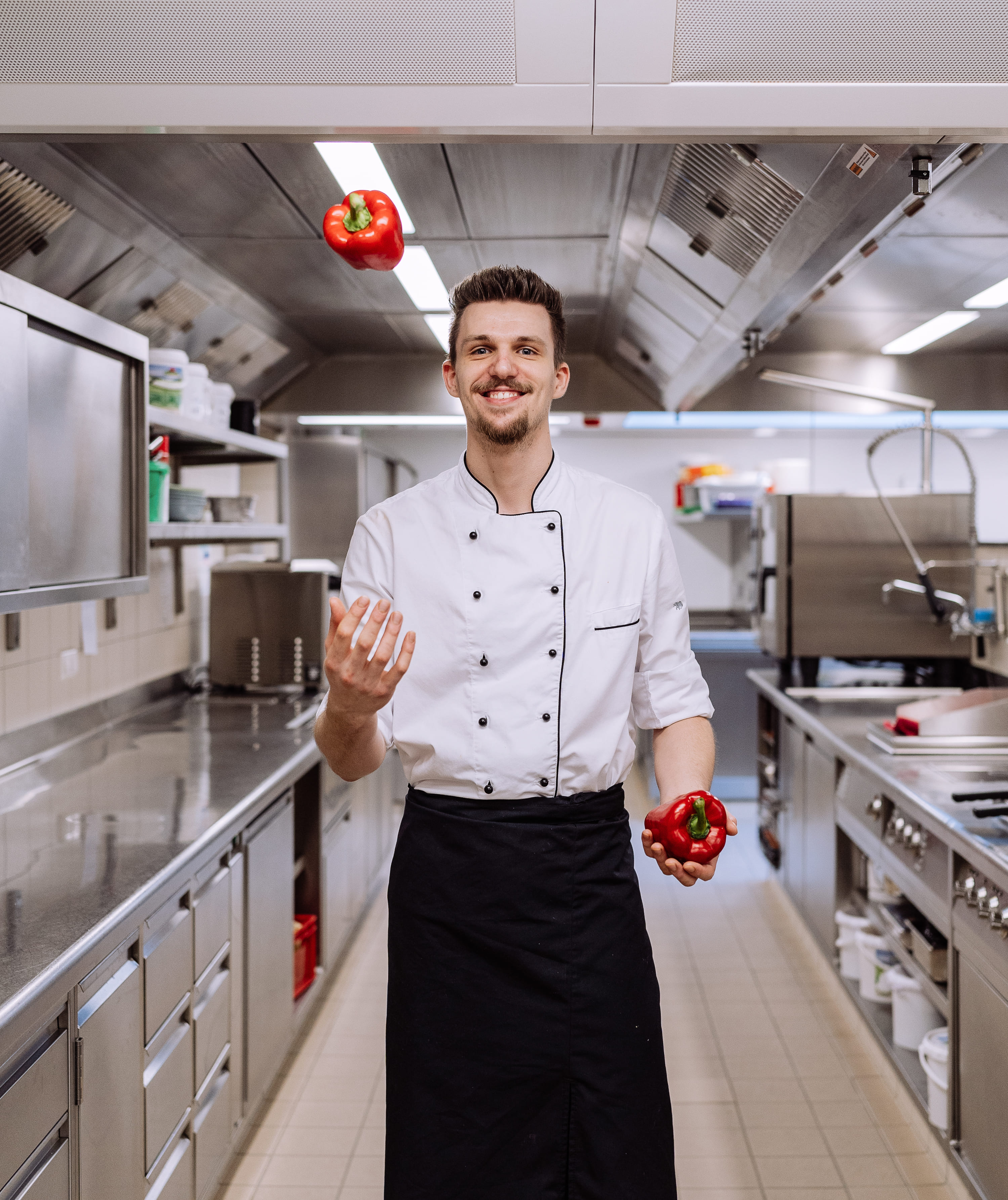 Koch jongliert in der Küche mit zwei roten Paprika.